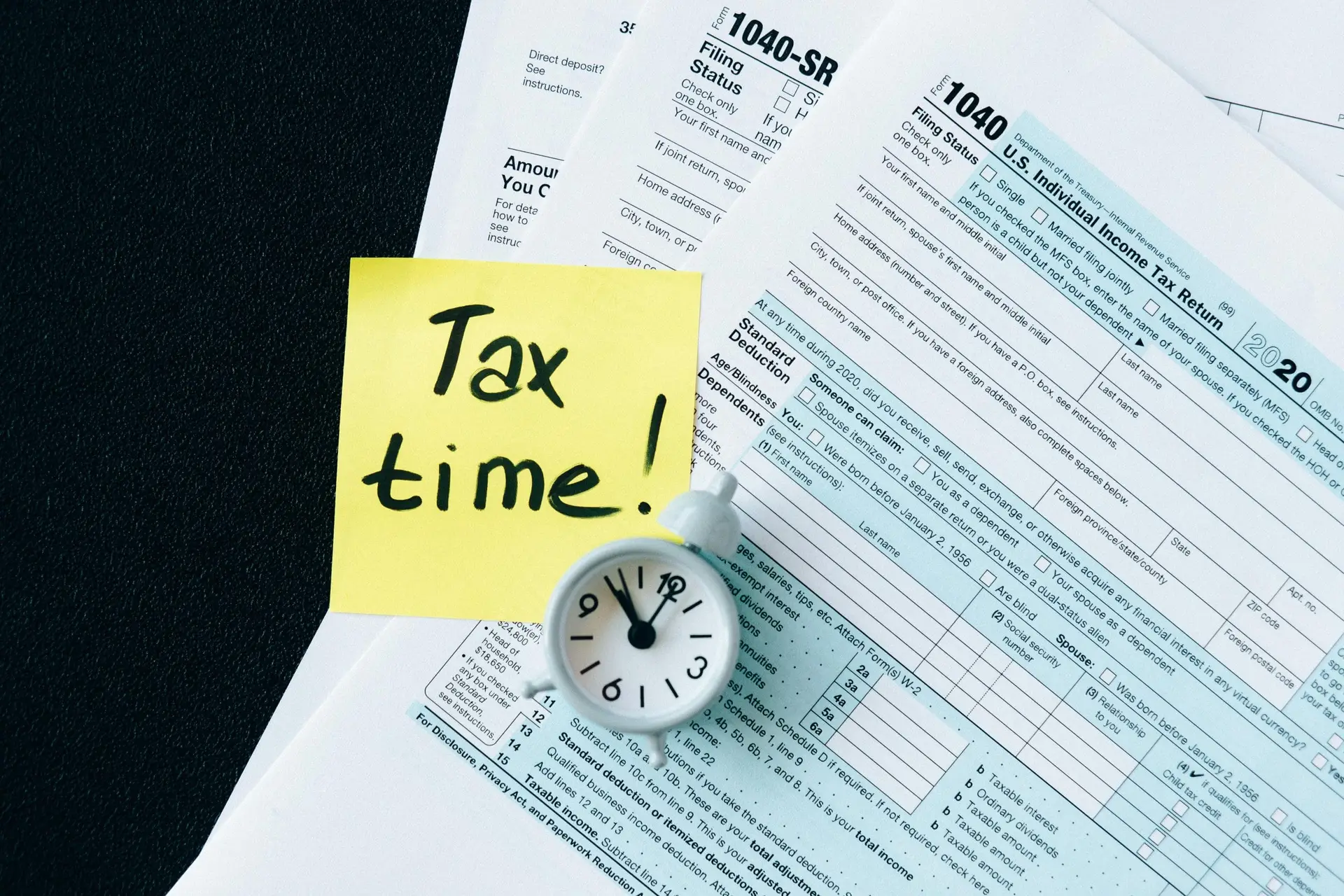 About Us A stack of tax forms with a clock and yellow sticky note saying 'Tax time!' indicating urgency.